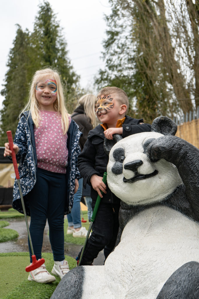 A young boy with a tiger face painting standing next to a girl wearing a purple jumper holding a red mini golf putter