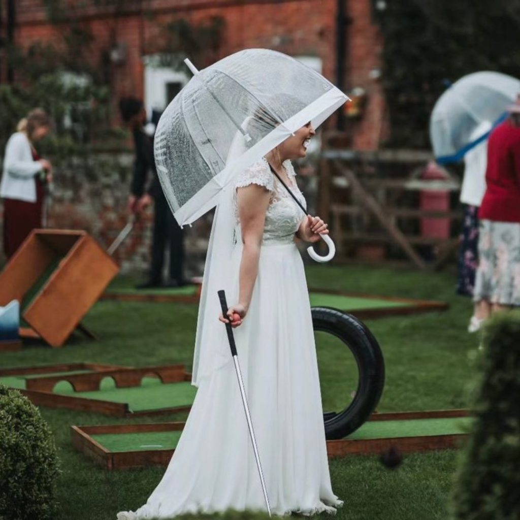 Bride wearing a wedding dress holding an umbrella in one hand and a mini golf putter in the other