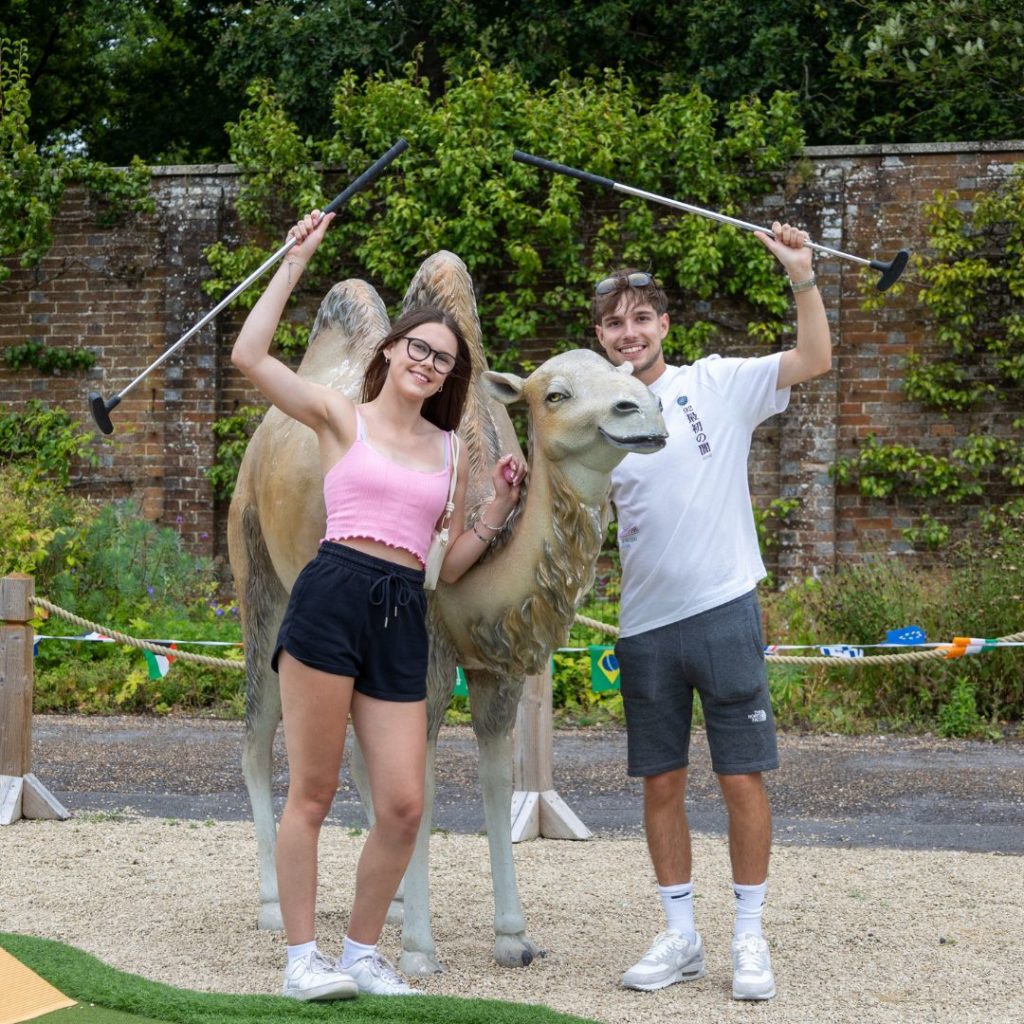 Girl and boy standing in between a lifesize camel prop with mini golf putters